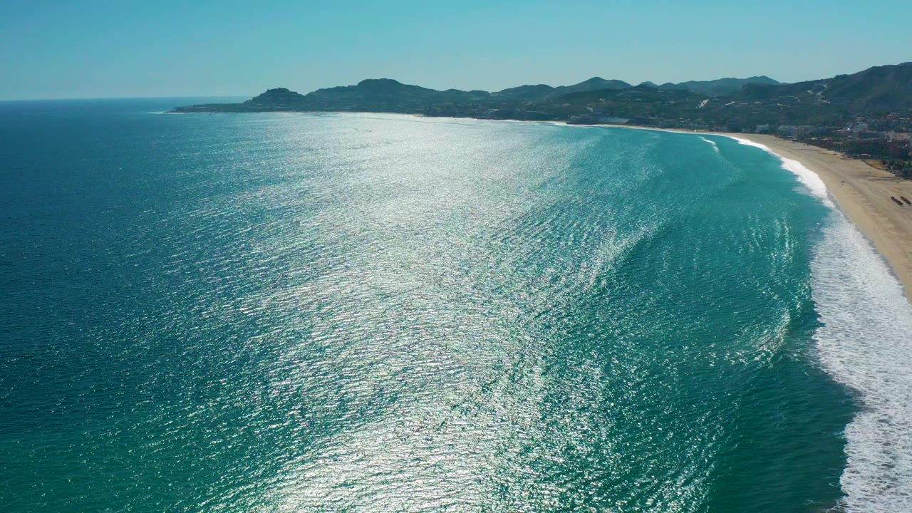 Aerial view over San Jose Del Cabo coastline with turquoise ocean, bright sun reflection, sandy beach at right, distant mountains on the horizon, and no visible people.
