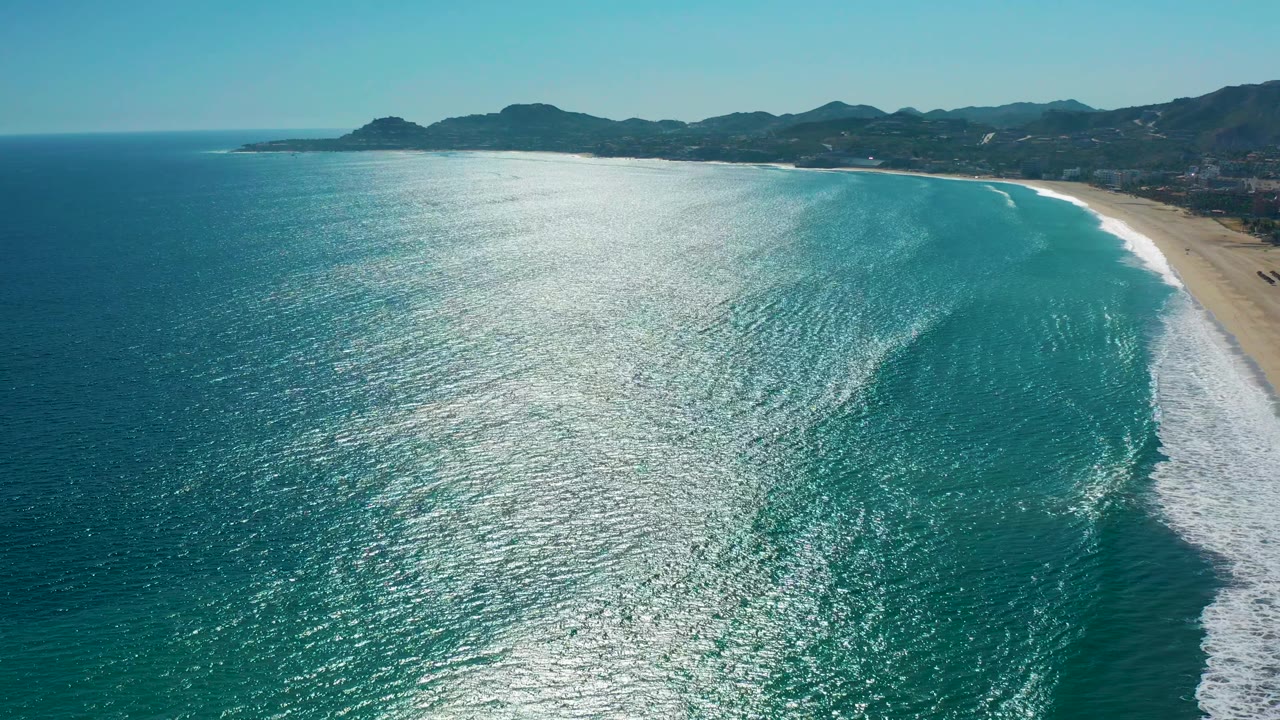 A wide aerial frame of San Jose Del Cabo coastline shows a large patch of sun glare over teal water, white wave foam near the beach, mountains inland, and no visible people.