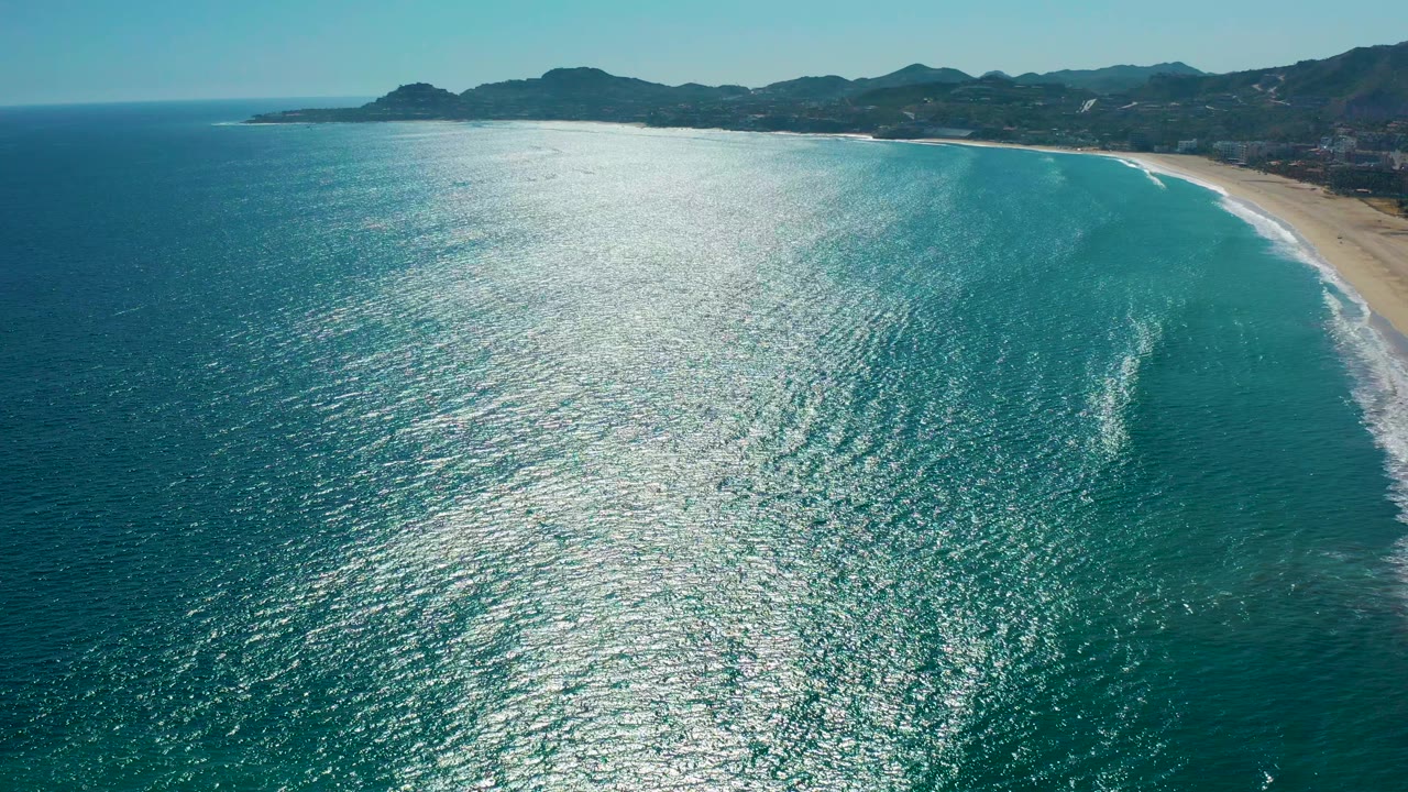 Aerial view over San Jose del Cabo turquoise water with a curved sandy beach on the right, distant mountains on the horizon, bright sun reflection on the ocean, and no visible people.