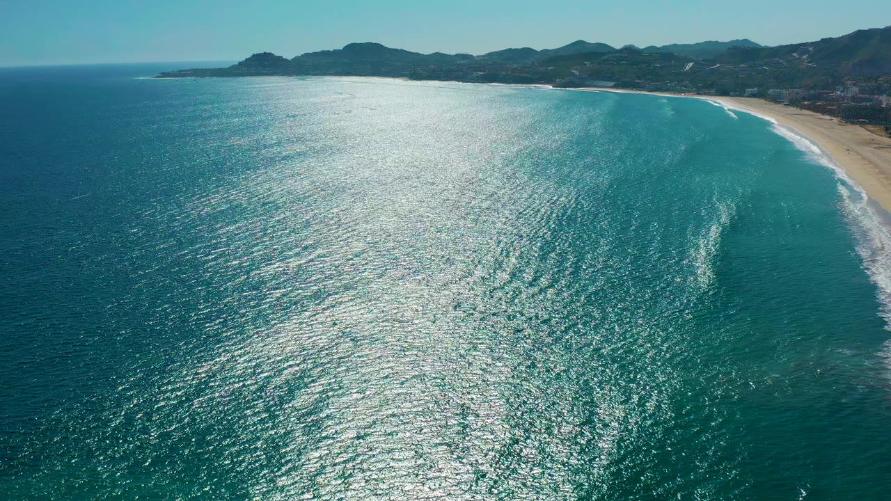 Sunlit ocean surface with waves approaching a sandy shoreline