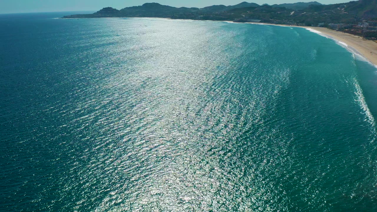 Aerial view over the San Jose del Cabo coast with sunlit turquoise water in the foreground, a sandy beach along the right edge, distant hills on the horizon, and no visible people.