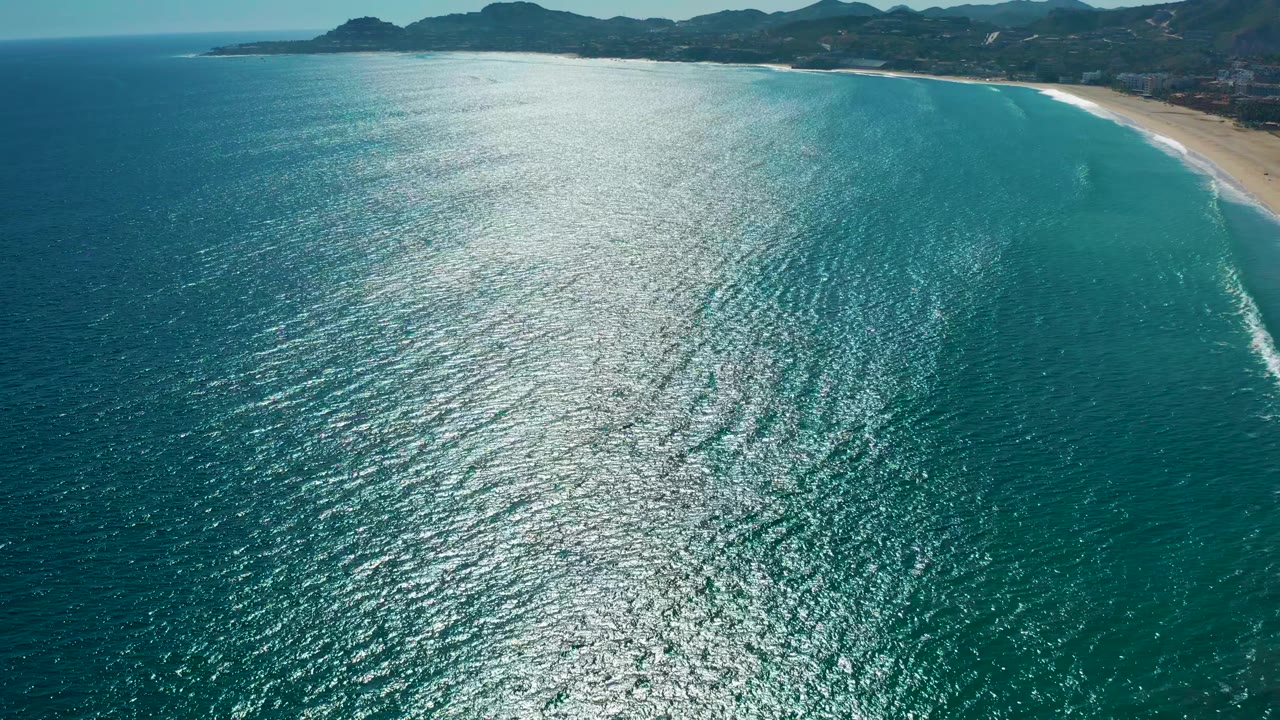 Wide aerial frame over blue-green water near San Jose del Cabo, showing small surf lines approaching the sandy beach, mountains across the top of frame, and no visible people.