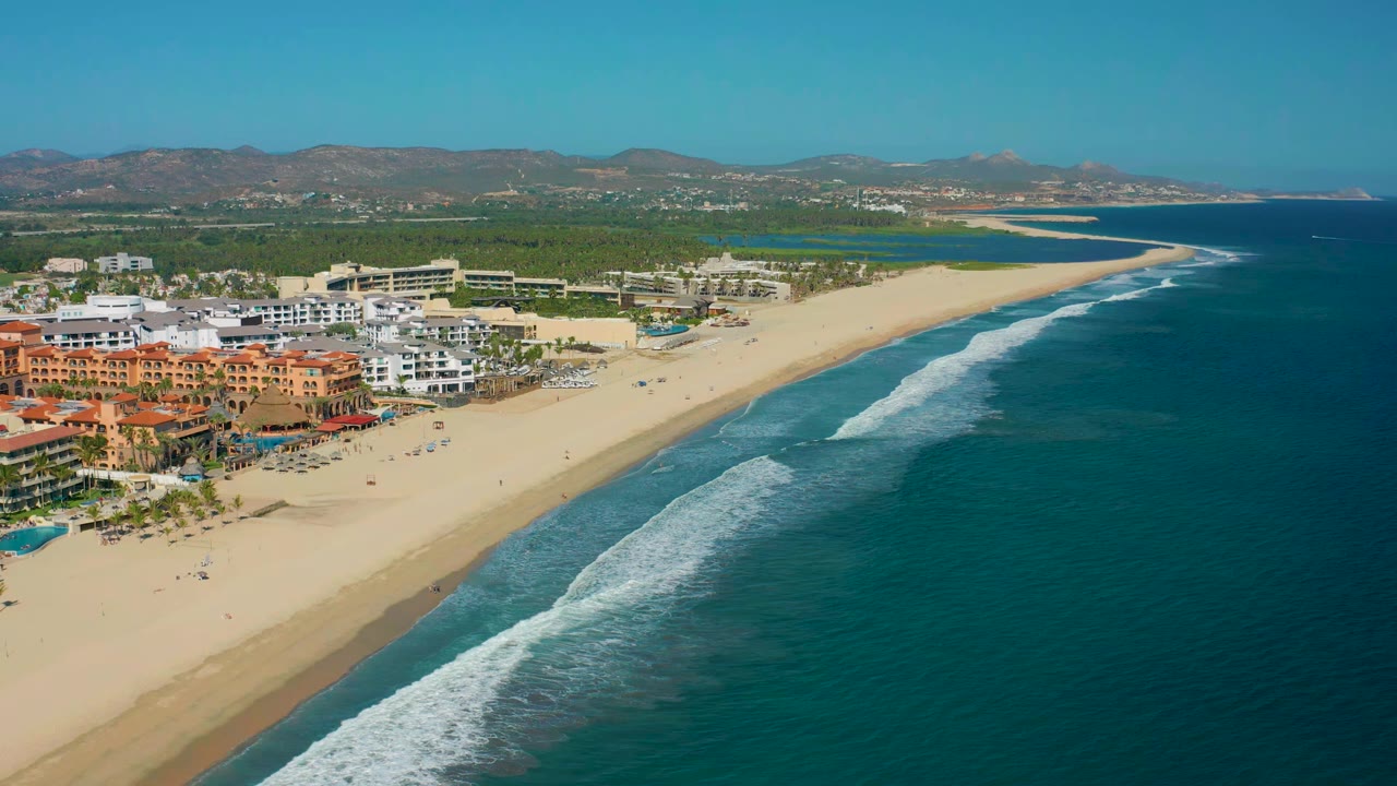 Early frame in the San Jose Del Cabo aerial pan, showing hotels clustered along the left side, a long curved beach, teal water, white wave bands, and no visible people.