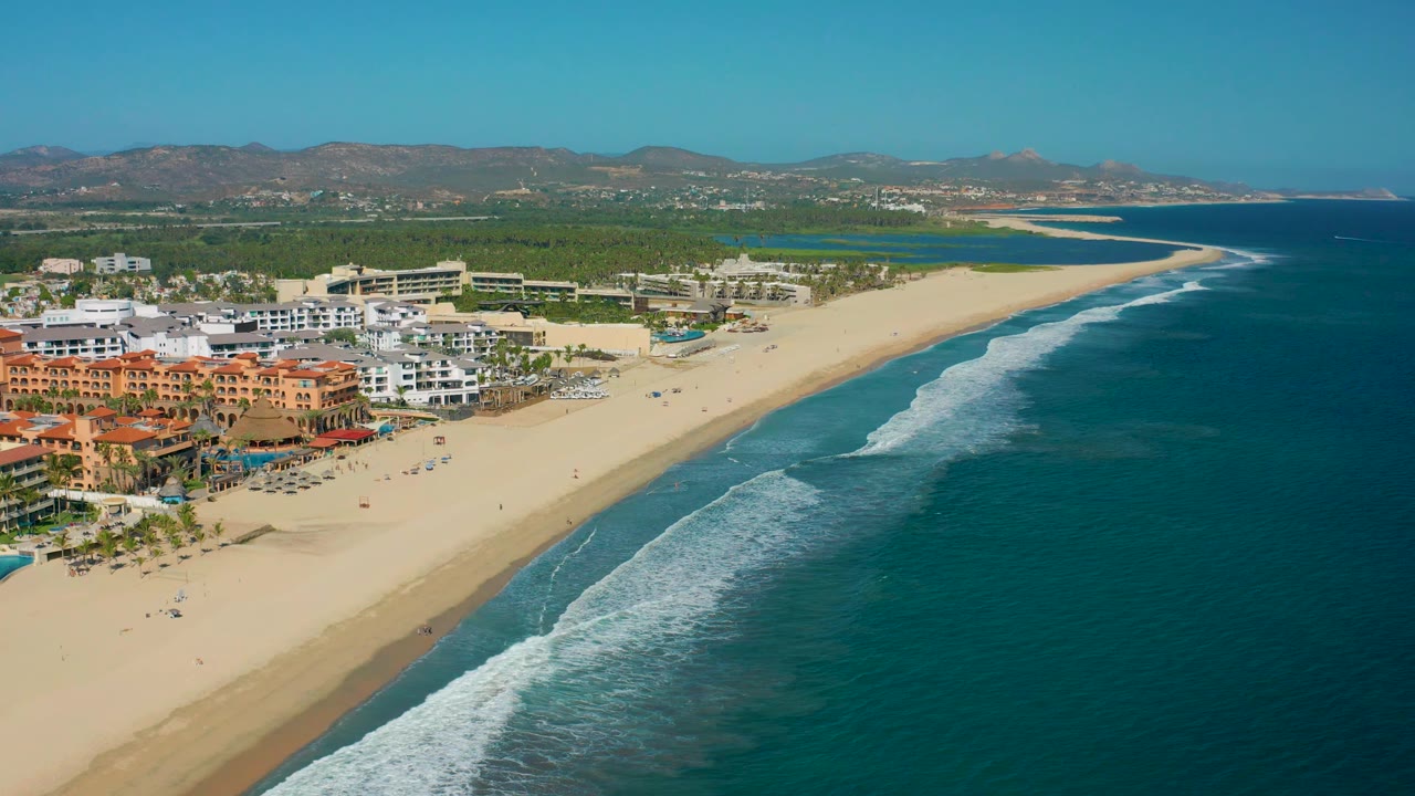 Mid-clip aerial frame over San Jose Del Cabo, with orange and white resort structures at lower left, broad sand, shallow surf texture near shore, mountains beyond, and no visible people.