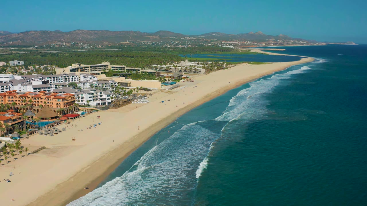 Wide aerial frame of San Jose del Cabo shoreline with white waves curling along the sand, ocean filling the right side, beachfront buildings on the left, and no visible people.