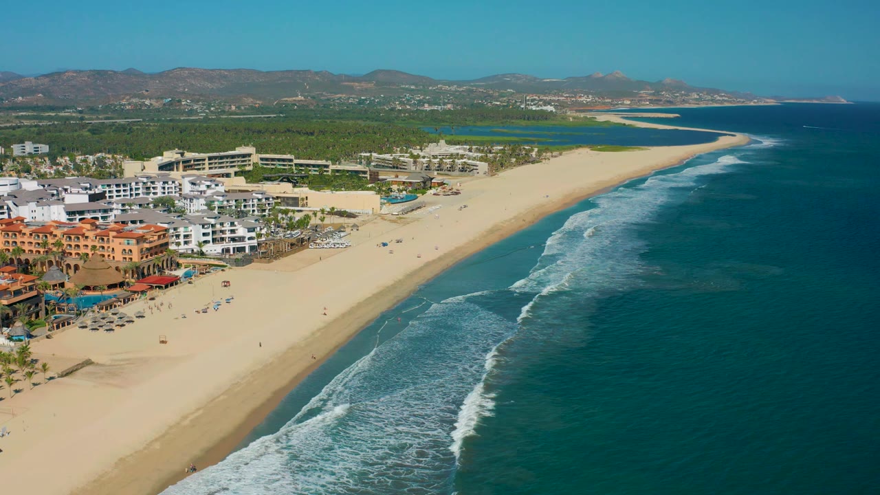 Aerial San Jose del Cabo beach view with surf lines advancing toward the sand, a lagoon-like water area behind the beach, clustered beachfront buildings, mountains, and no visible people.