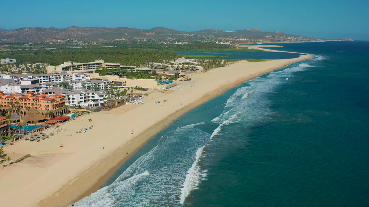 Aerial coastal view with sandy beach, rolling surf, hotels, greenery, and mountains.