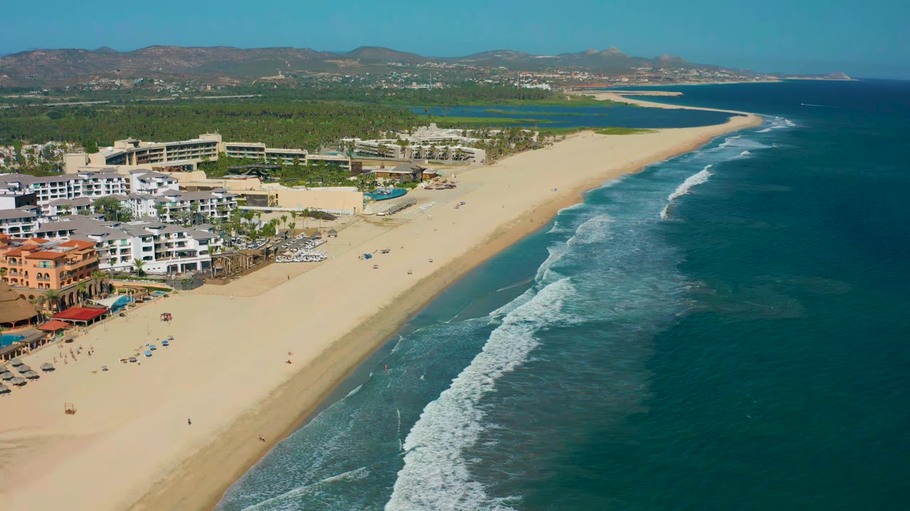 Drone view over San Jose del Cabo with the shoreline running diagonally, waves breaking near shore, resort buildings and pools on the left, and distant hills beyond green inland land.