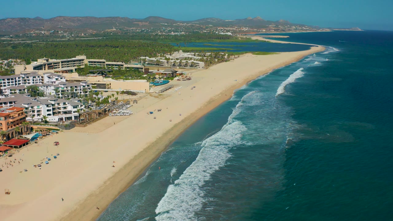 Aerial coastline view with resort pools, beach umbrellas, surf, ocean, and hills.