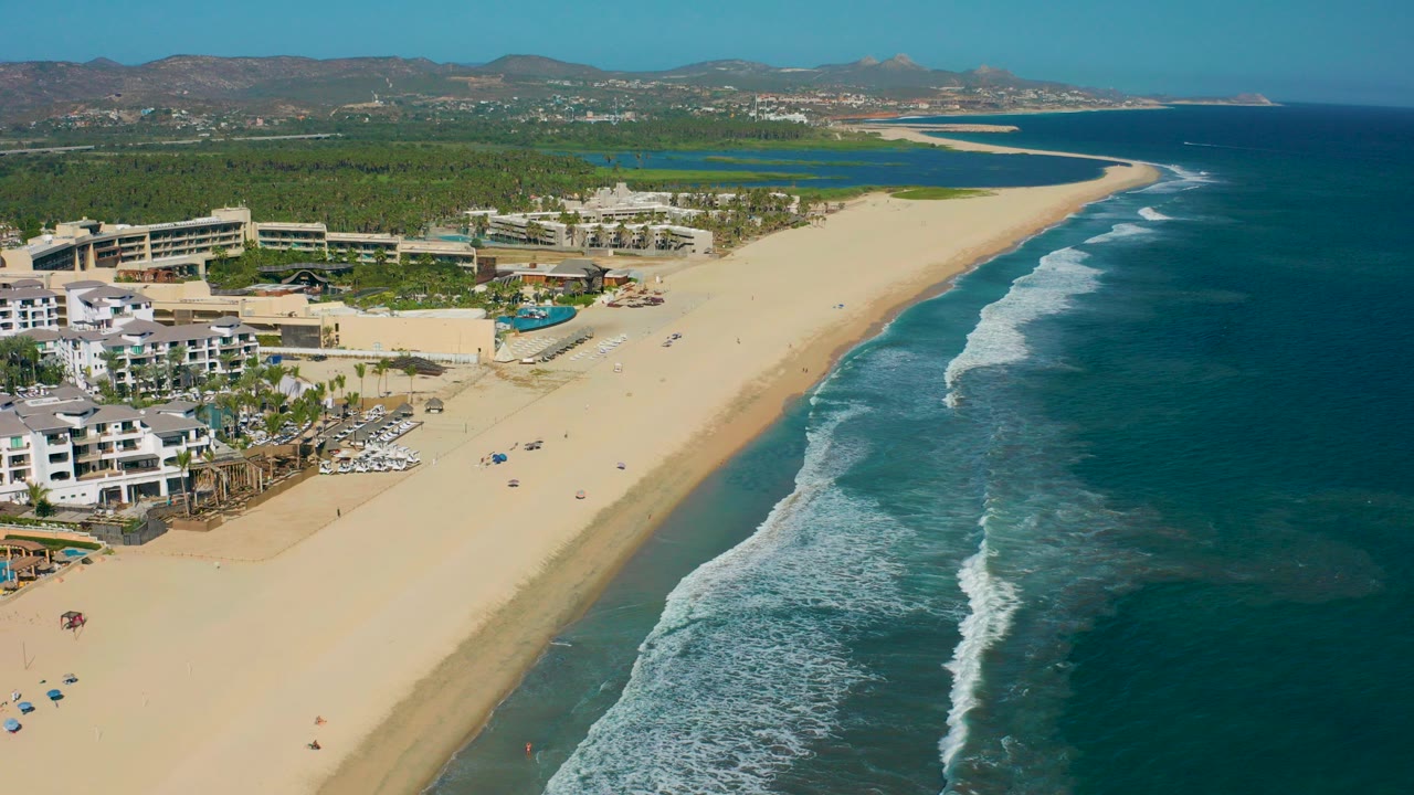 High aerial view in San Jose del Cabo, Mexico showing resort buildings and a blue pool at left, a wide sandy beach through the center, and white surf lines along the ocean at right.
