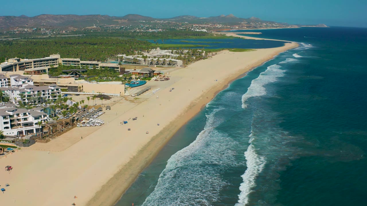 San Jose del Cabo aerial view with the resort complex shifted toward the left edge, a long open beach running toward the background, and deeper blue ocean filling the right side.