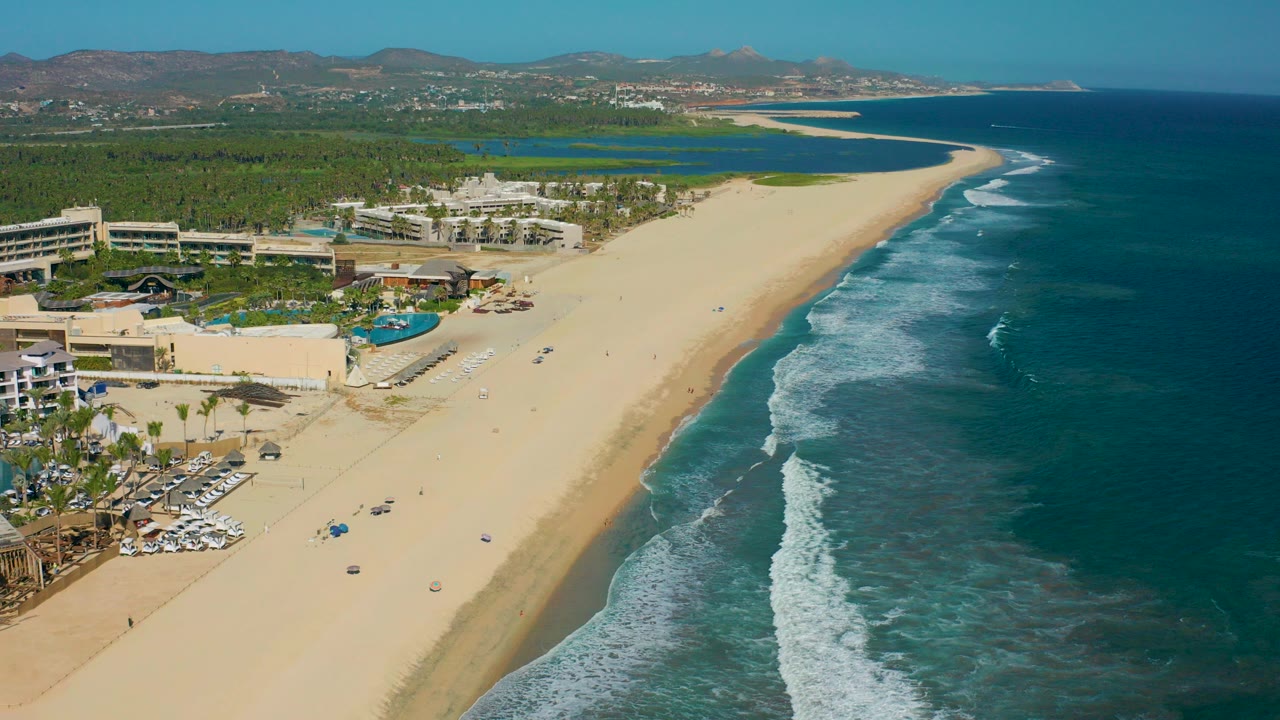Opening aerial frame over San Jose del Cabo beach with resort buildings and pools at left, wide sand in the center, ocean surf at right, distant hills beyond, and no visible people.