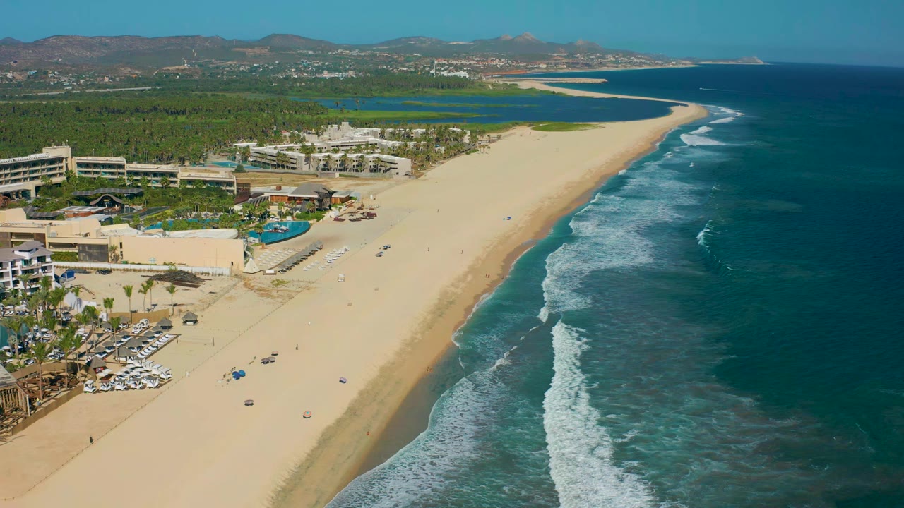 Wide aerial coastline with resort pools, palm trees, sand beach, and rolling waves.