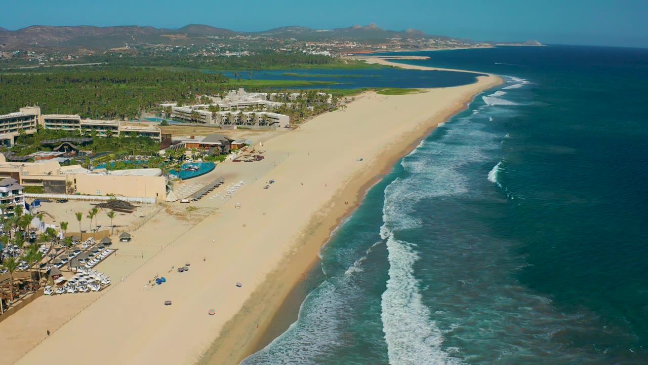 San Jose del Cabo beach remains centered under bright light, with the resort complex and pool deck at left, deeper blue ocean at right, and no visible people.