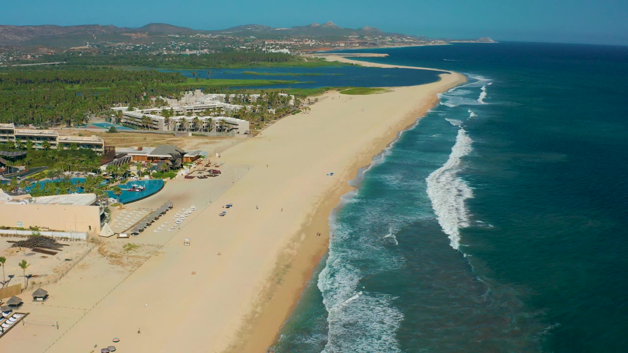 Aerial frame over San Jose del Cabo showing resort pools at lower left, a broad sandy beach through the center, blue ocean surf on the right, and distant hills beyond the palms.