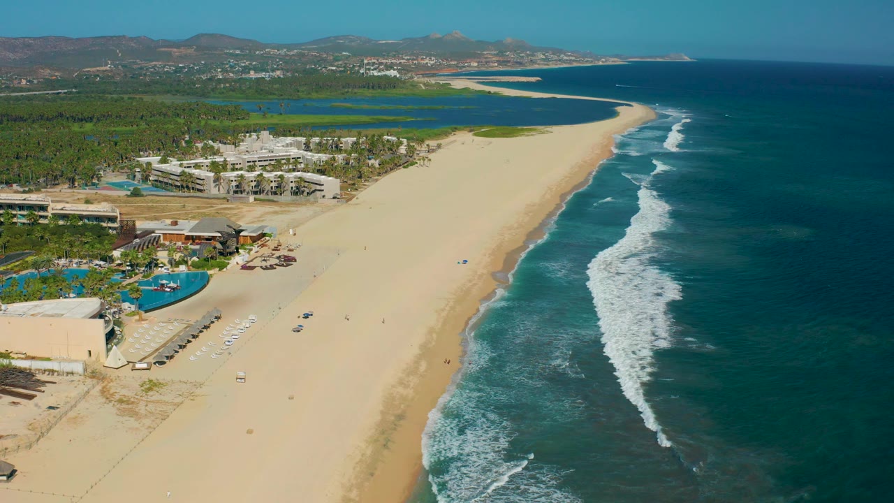 Wide aerial beach view with hotel buildings, pools, palms, and surf line.