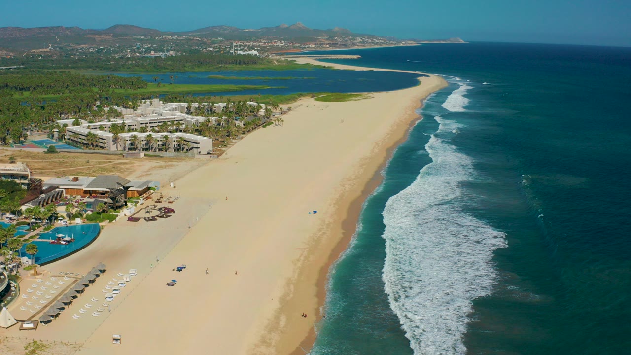Aerial view over San Jose del Cabo with resort buildings and pools at lower left, a wide sandy beach through the center, blue ocean surf at right, and small beachgoers visible on the sand.