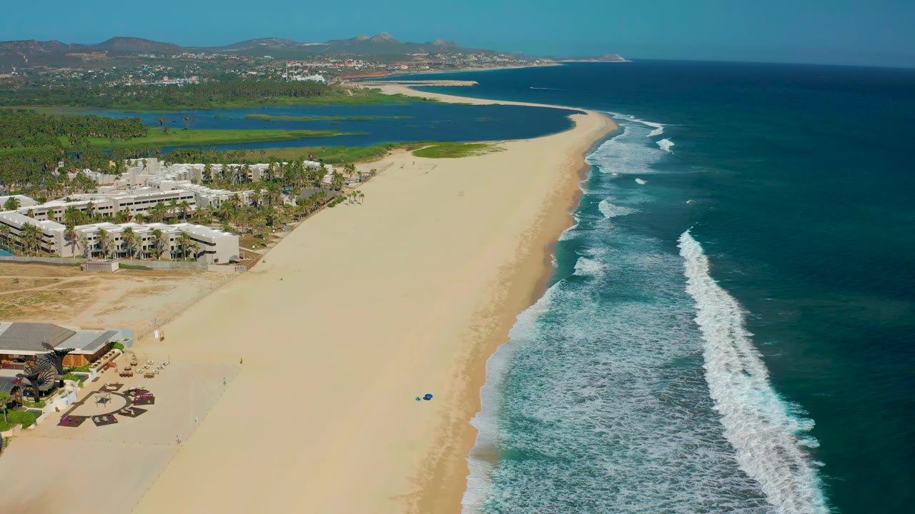 San Jose Del Cabo coastline from above, with the surf line slightly farther down the beach, resort buildings at lower left, green inland water behind the palms, and no visible people.