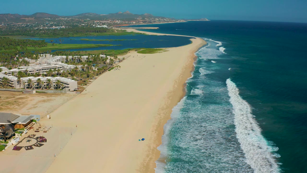 Aerial San Jose Del Cabo beach view with brighter surf foam on the right, open sand in the middle, low coastal structures at left, green lagoons inland, and no visible people.