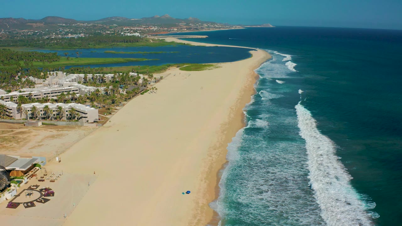 San Jose Del Cabo shoreline seen from an elevated angle, with white surf running along the right edge, broad sand in the center, resort buildings at lower left, and no visible people.