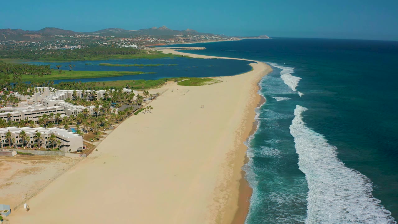 San Jose del Cabo coastline seen from above with pale sand curving past resort buildings, a green-blue lagoon inland, foamy surf along the right edge, distant hills, and no visible people.