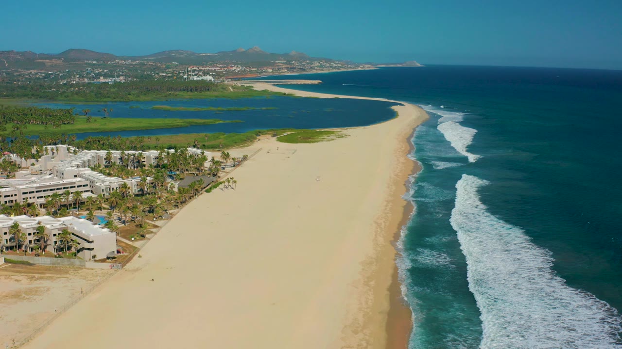 San Jose del Cabo aerial coastline with resort buildings clustered at lower left, a mostly empty sandy beach through the center, a dark blue surf band on the right, lagoon at upper left, and no visible people.