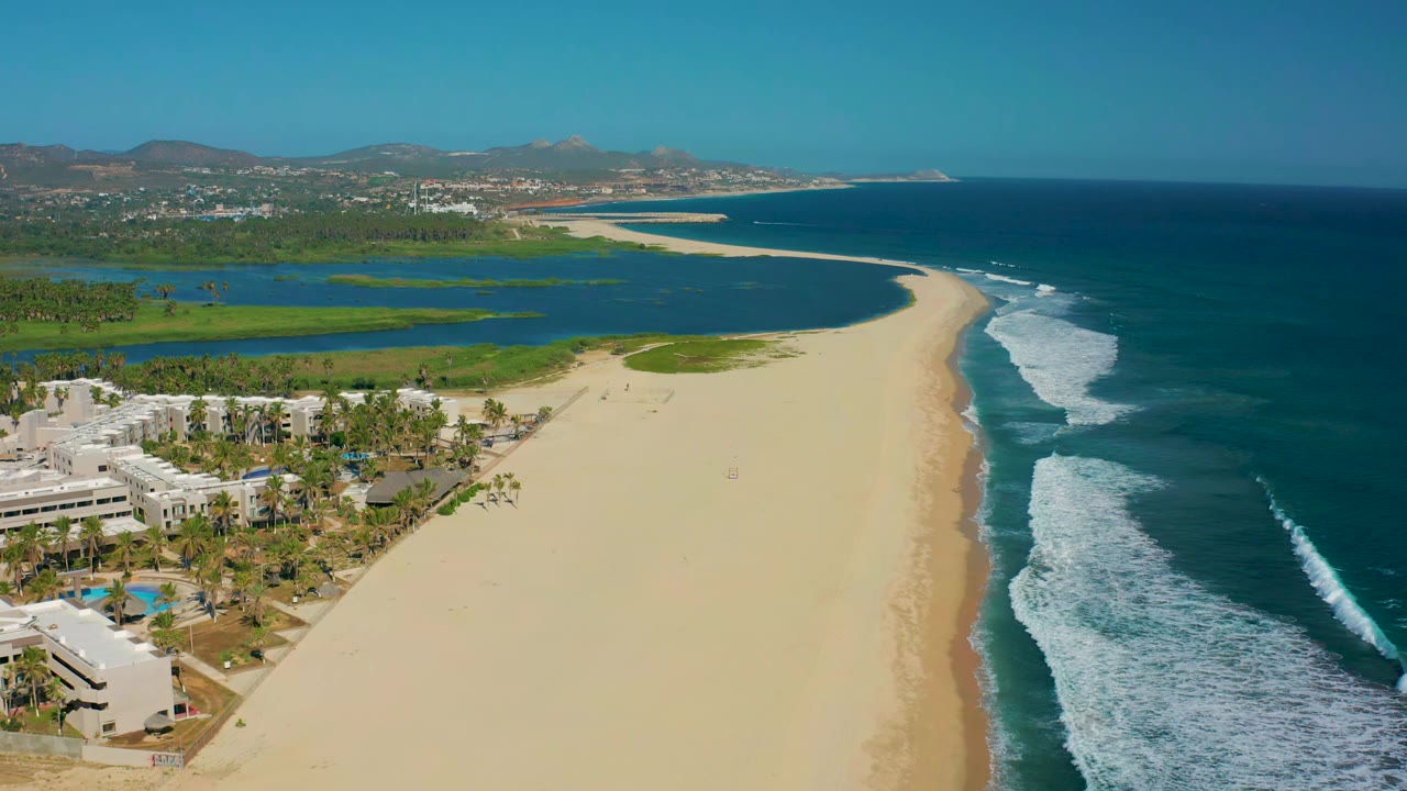 High aerial frame over San Jose Del Cabo with pale sand centered, lagoon and green vegetation to the left, ocean surf to the right, distant hills on the horizon, and no visible people.