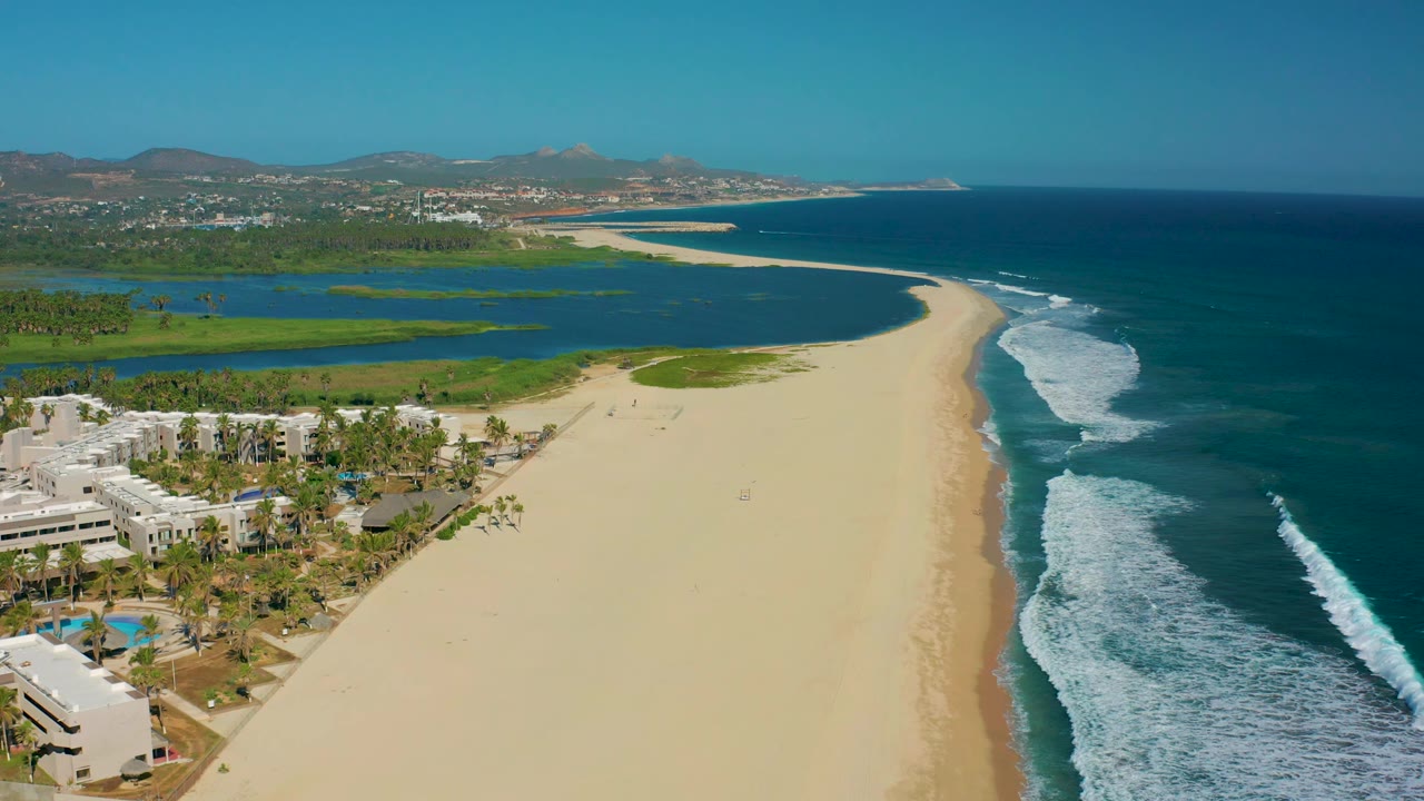 Aerial view of the San Jose Del Cabo shoreline with resort-style white buildings and palms at lower left, a broad empty beach in the middle, lagoon water inland, and rolling ocean waves on the right.