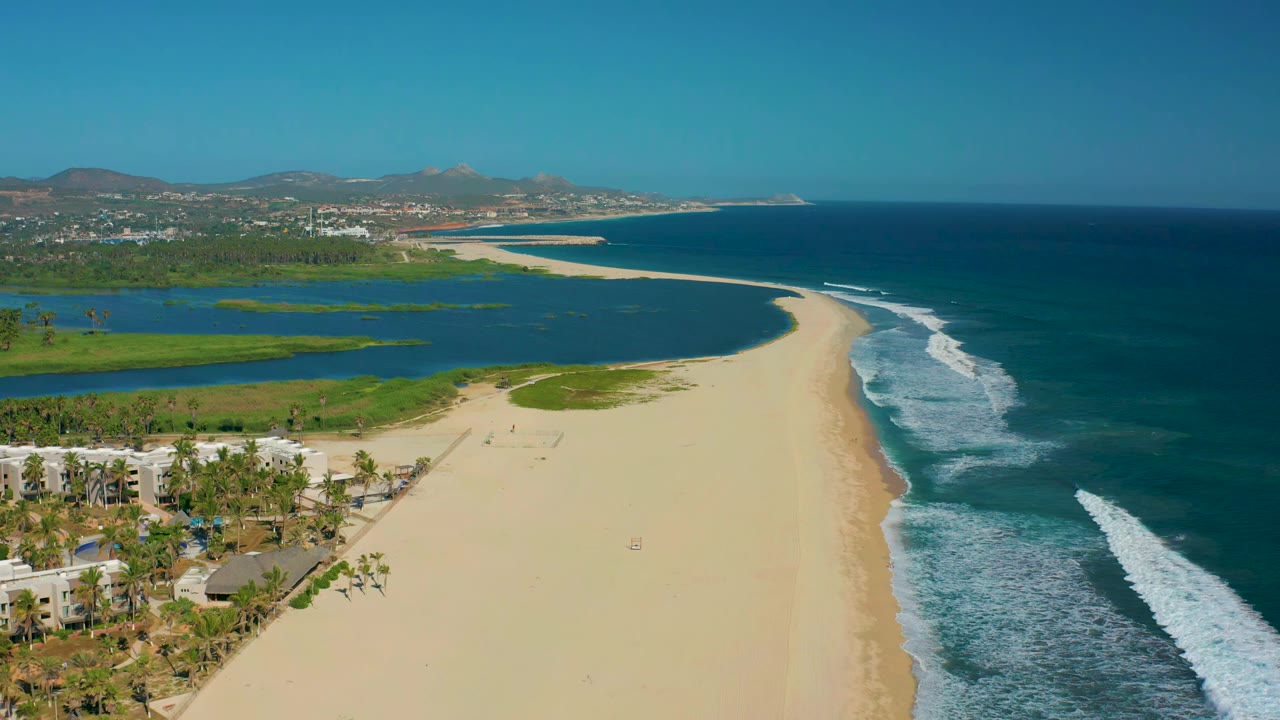 Early aerial frame over San Jose del Cabo, Mexico showing the sandy shoreline stretching ahead, green lagoon water to the left, blue ocean to the right, and no visible people.
