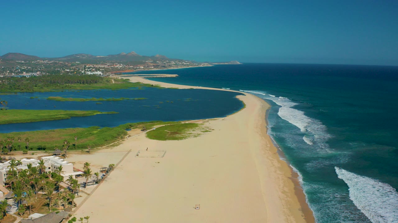 Opening aerial frame over San Jose del Cabo showing a wide sandy beach between a green-blue lagoon on the left and ocean waves on the right, with palm trees and buildings at the lower left and no visible people.