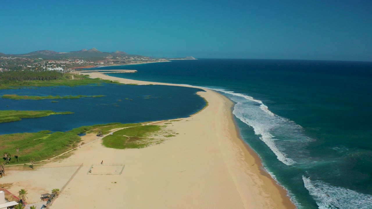 Aerial view in San Jose Del Cabo, Mexico, with a wide sandy beach between a lagoon and breaking ocean waves, with tiny people visible on the sand near the lower left.