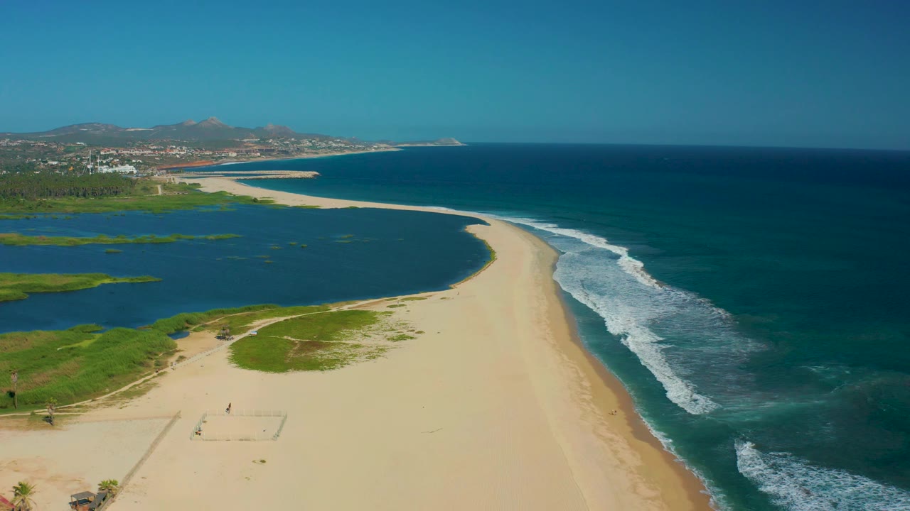 Aerial view over San Jose Del Cabo beach with broad empty sand, a lagoon along the left edge, ocean waves to the right, and tiny people visible near the lower-left beach area.