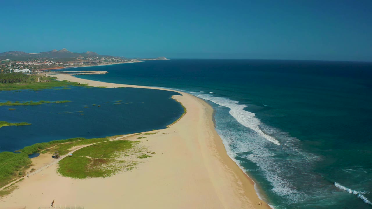 High aerial frame over San Jose del Cabo, Mexico, with the curving beach centered between dark lagoon water on the left and white wave lines on the ocean side, no visible people.