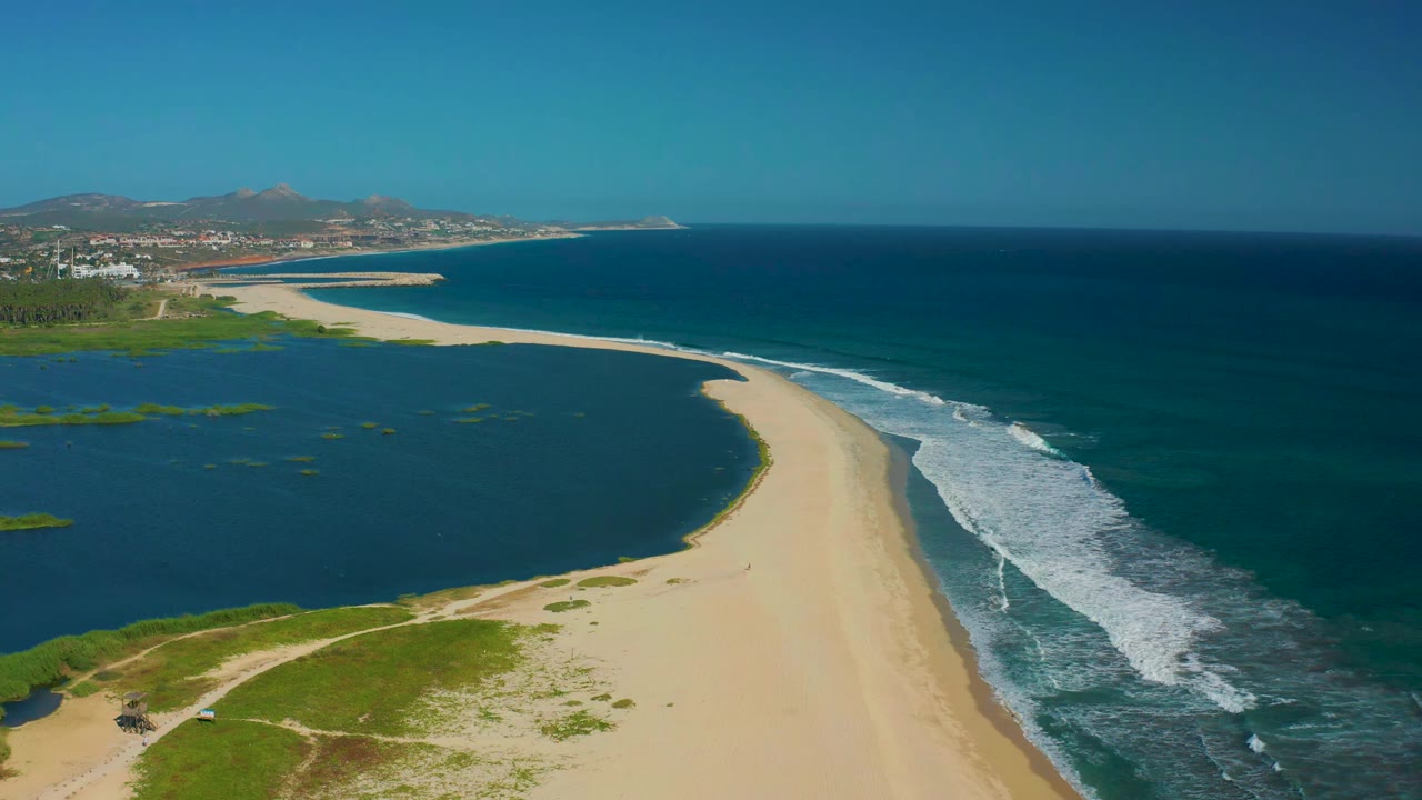 Aerial frame of the San Jose del Cabo sandbar curving toward distant shoreline buildings, with lagoon water left, ocean surf right, and no visible people.