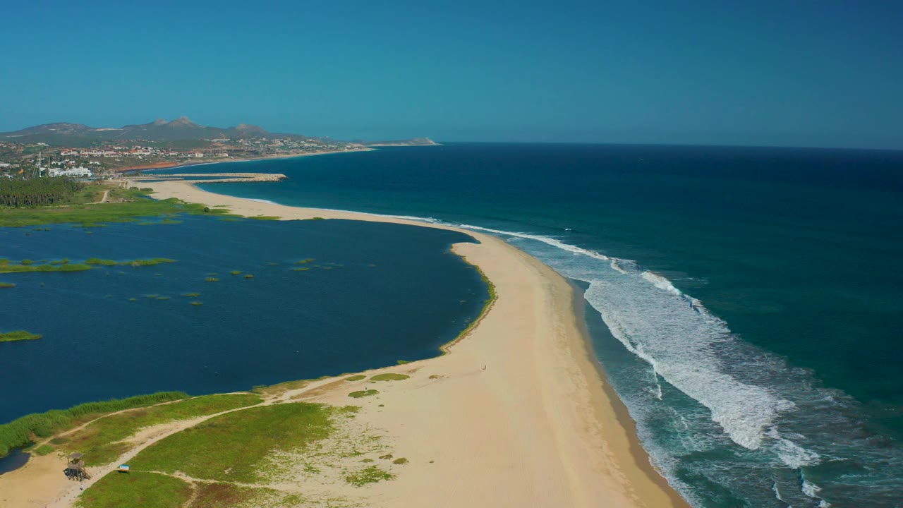 Wide aerial view over San Jose del Cabo coastline, with the sandbar running through the center, lagoon patches on the left, ocean waves on the right, and no visible people.