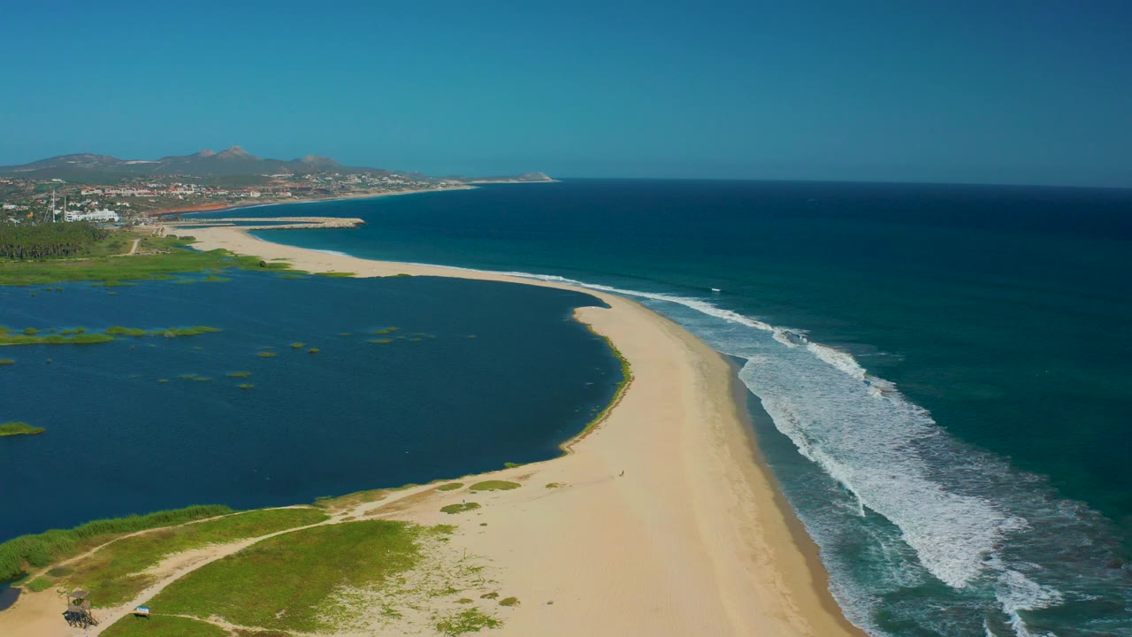 Aerial view in San Jose del Cabo with lagoon water filling the left side, a sandy strip through the center, ocean surf along the right edge, and no visible people.