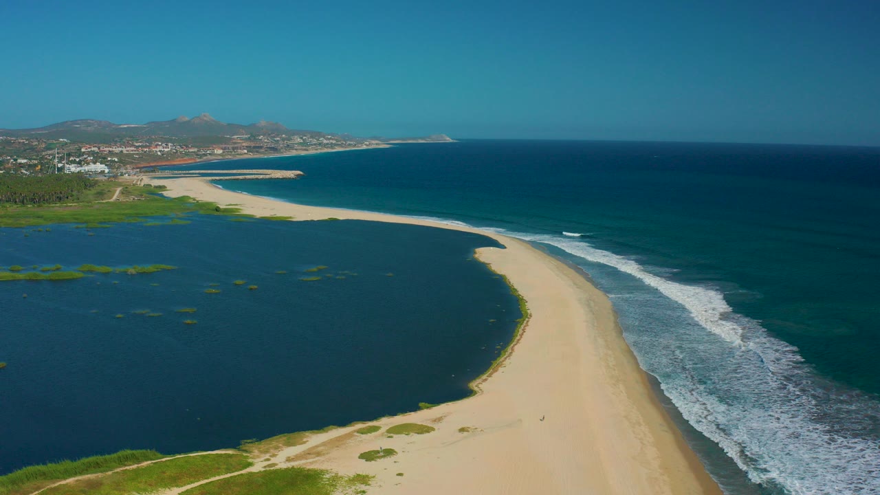 Aerial view in San Jose del Cabo of a pale sandy spit separating dark lagoon water from blue ocean surf, with distant buildings and hills on the horizon.