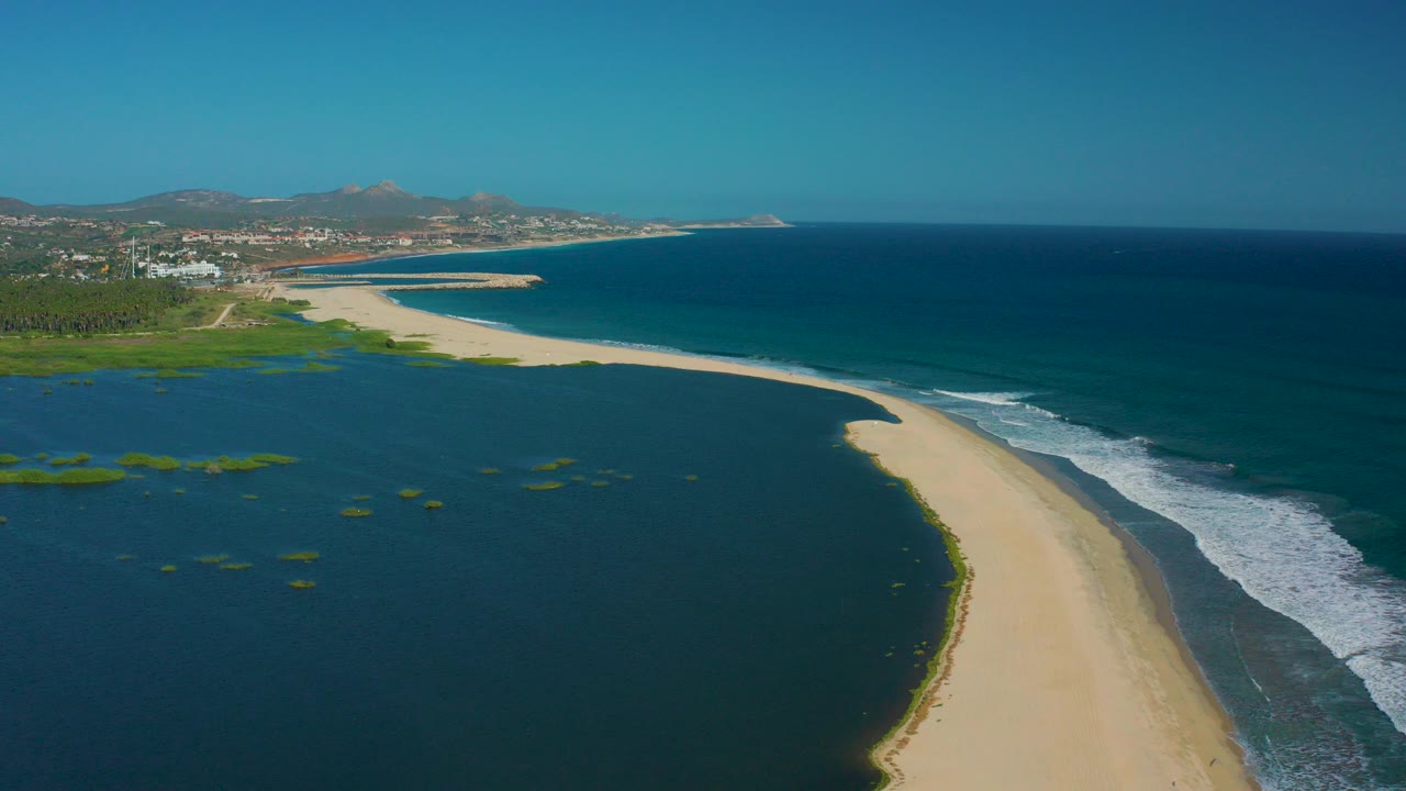 Aerial view in San Jose Del Cabo, Mexico, showing a curved sandbar between dark lagoon water and blue ocean surf, with distant hills and no visible people.