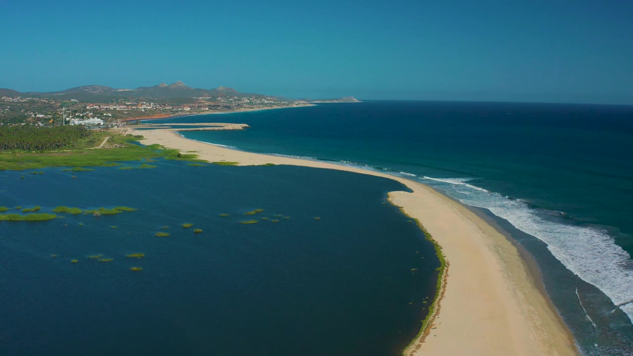 Aerial view of the San Jose Del Cabo sand curve with calm lagoon water on one side, ocean surf on the other, hills in the distance, and no visible people.