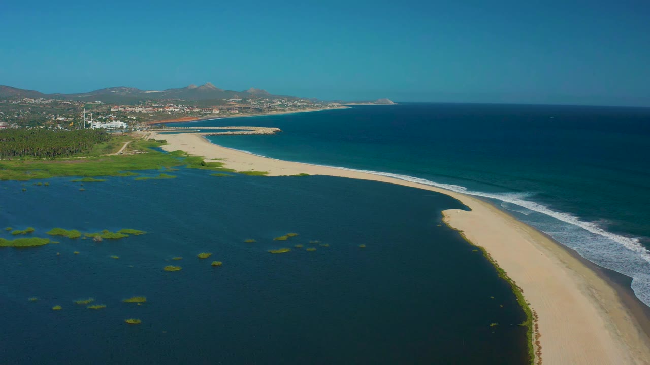 Aerial coastal view with lagoon foreground, sand spit, surf, hills, and buildings.
