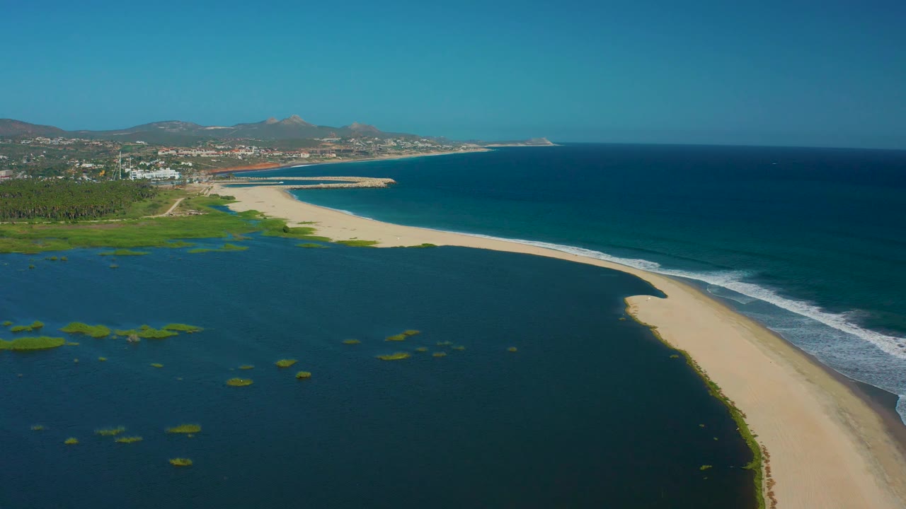 Aerial view in San Jose del Cabo with green lagoon patches in dark water, a bright sandbar, white surf along the ocean side, distant town buildings, hazy hills, and no visible people.