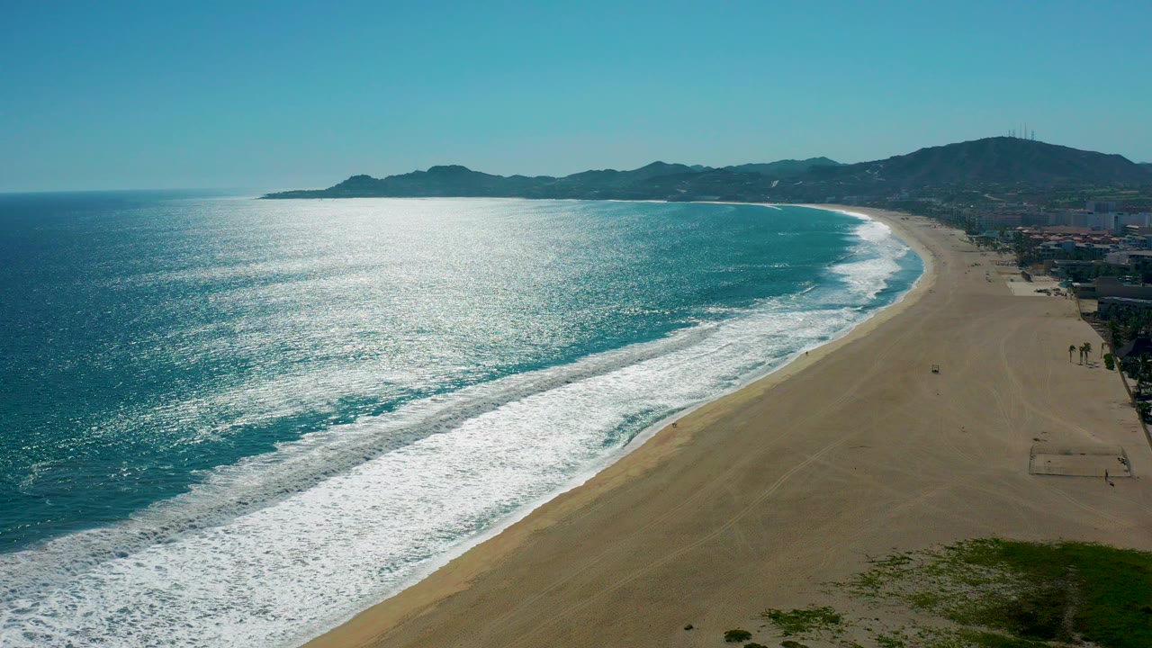 Aerial view of San Jose del Cabo coastline with turquoise water, white surf, a broad sandy beach, distant mountains, and tiny people on the right side of the shore.