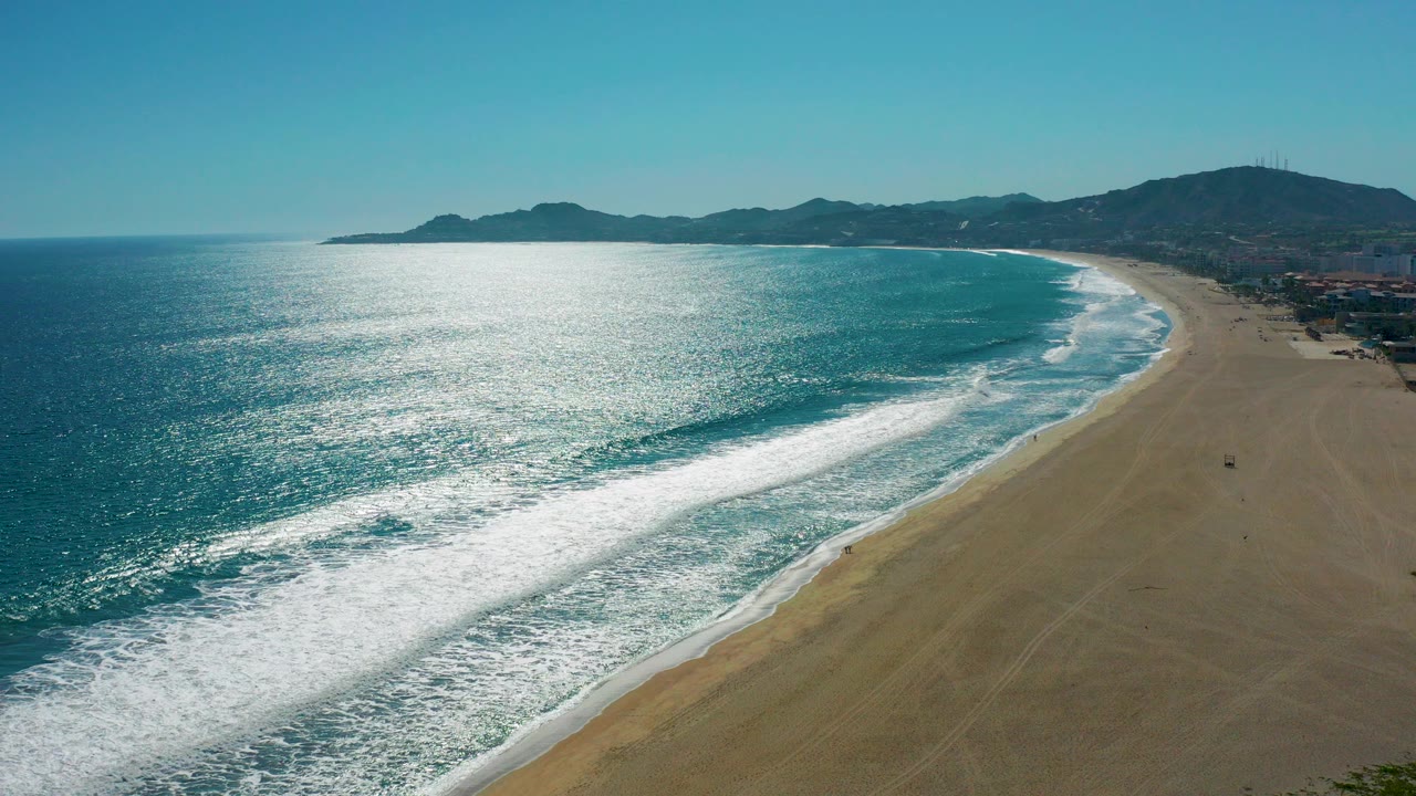 San Jose del Cabo coastline from above, with bright ocean glare on the left, white surf crossing the sand, distant hills on the horizon, and no visible people in this frame.