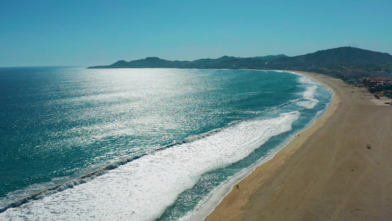 Wide aerial coastline with bright surf bands rolling toward a long sandy shore.