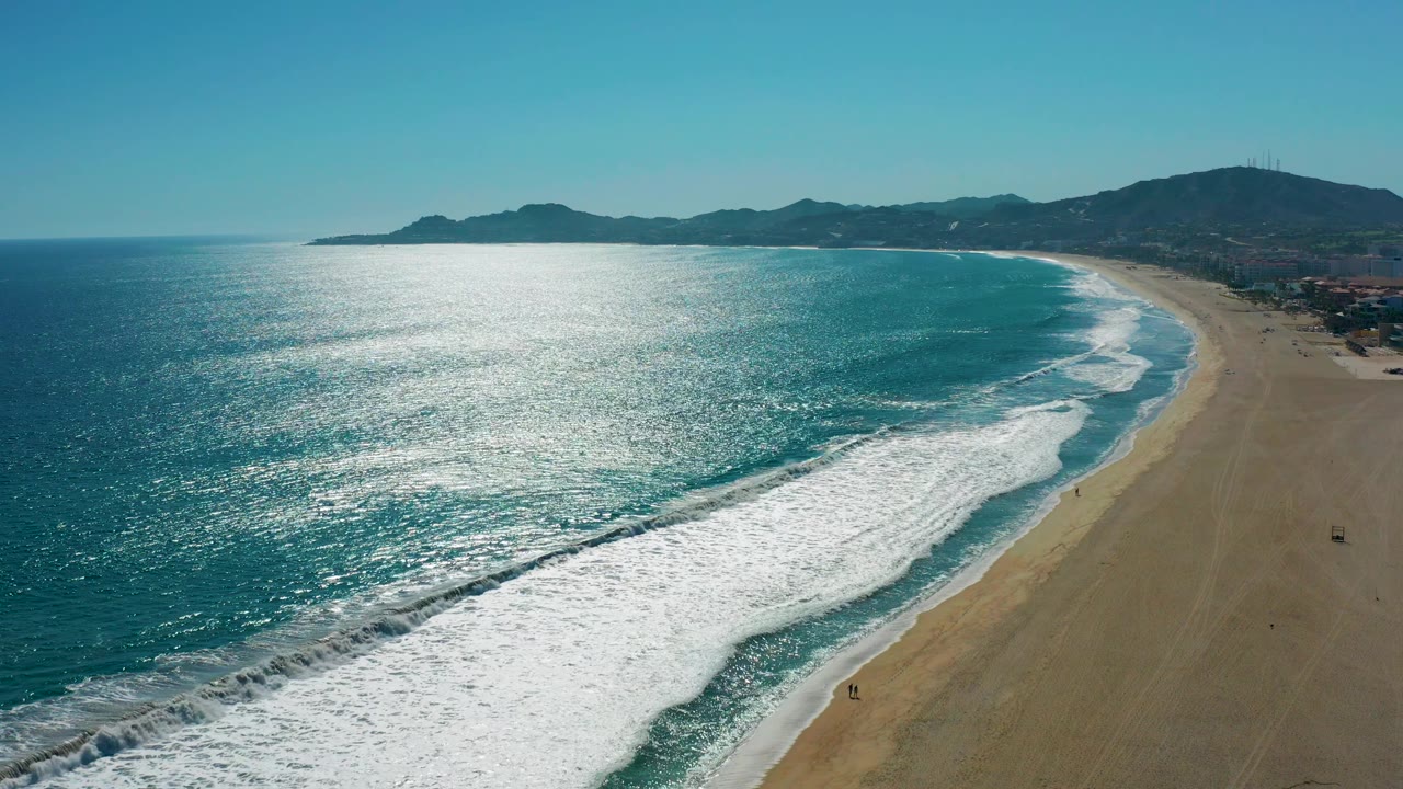 Aerial view in San Jose del Cabo with a large foamy wave spreading diagonally across the lower frame, open blue water to the left, empty-looking sand to the right, distant beachfront buildings, and mountains behind.