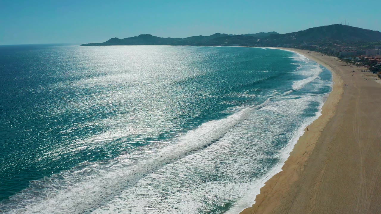 Aerial view in San Jose del Cabo, Mexico with shimmering blue-green water, foam lines moving toward the sand, distant mountains, and no visible people.