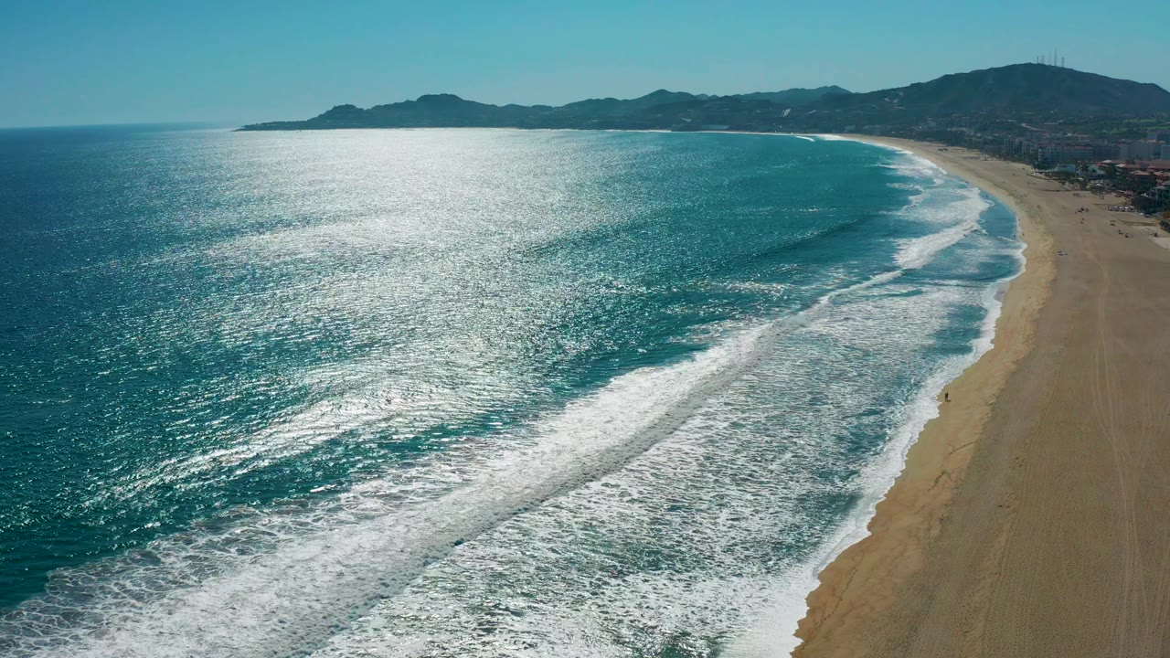 San Jose del Cabo, Mexico coastline seen from above, with a broad band of foamy surf near the beach, darker water offshore, hillside background, and no visible people.
