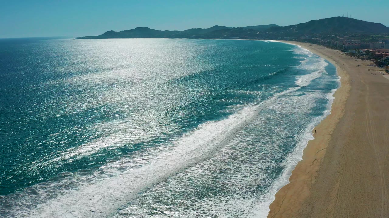 A wide aerial frame over San Jose del Cabo, Mexico shows sparkling ocean water, layered white waves, a long sandy beach, distant hills, and no visible people.