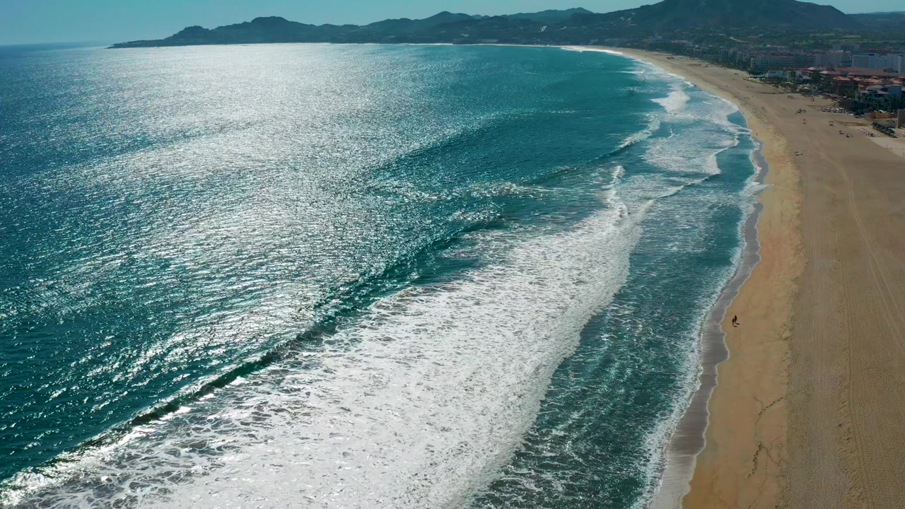 Aerial frame of San Jose Del Cabo shoreline with shimmering water at left, a broken wave line in the center, broad sand at right, distant hills, and a tiny walker visible near the surf edge.
