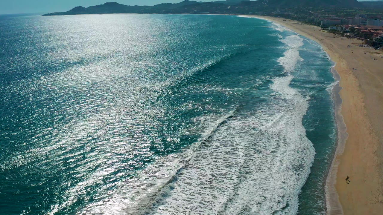 Sunlit ocean waves break beside a long sandy beach with mountains beyond.